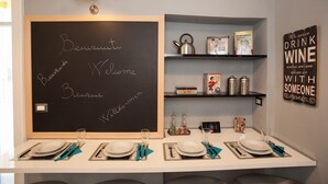 A neatly arranged dining area with a chalkboard displaying "Bienvenue Welcome" in multiple languages, a shelf with kitchen items, and a sign saying "We never drink wine unless we are alone or with someone." - Largodonnaregina home vacation home 2 bedroom apartment historic center (Napoli)