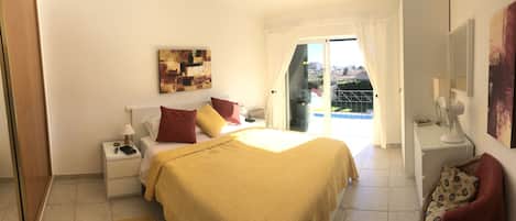 Une chambre à coucher avec un lit, des tables de chevet, une fenêtre avec des rideaux, un tableau au mur et une vue sur le balcon.