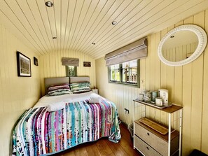 Ein Schlafzimmer mit Bett, Nachttisch mit Spiegel, Fenster mit Jalousien und einer kleinen Ablagefläche mit Gegenständen.