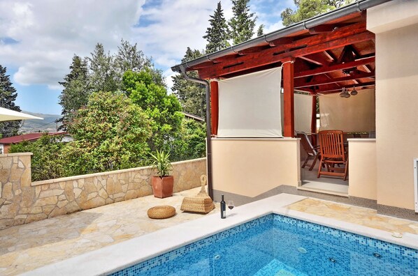 A swimming pool with stone tiles, a pergola with red beams, and a view of trees and a house in the background.
