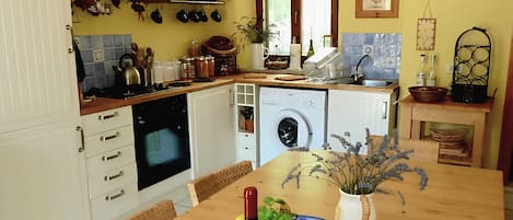 A compact kitchen with white cabinets, a built-in oven, a washing machine, and a dining table with a bowl of fruit and a bottle of wine.