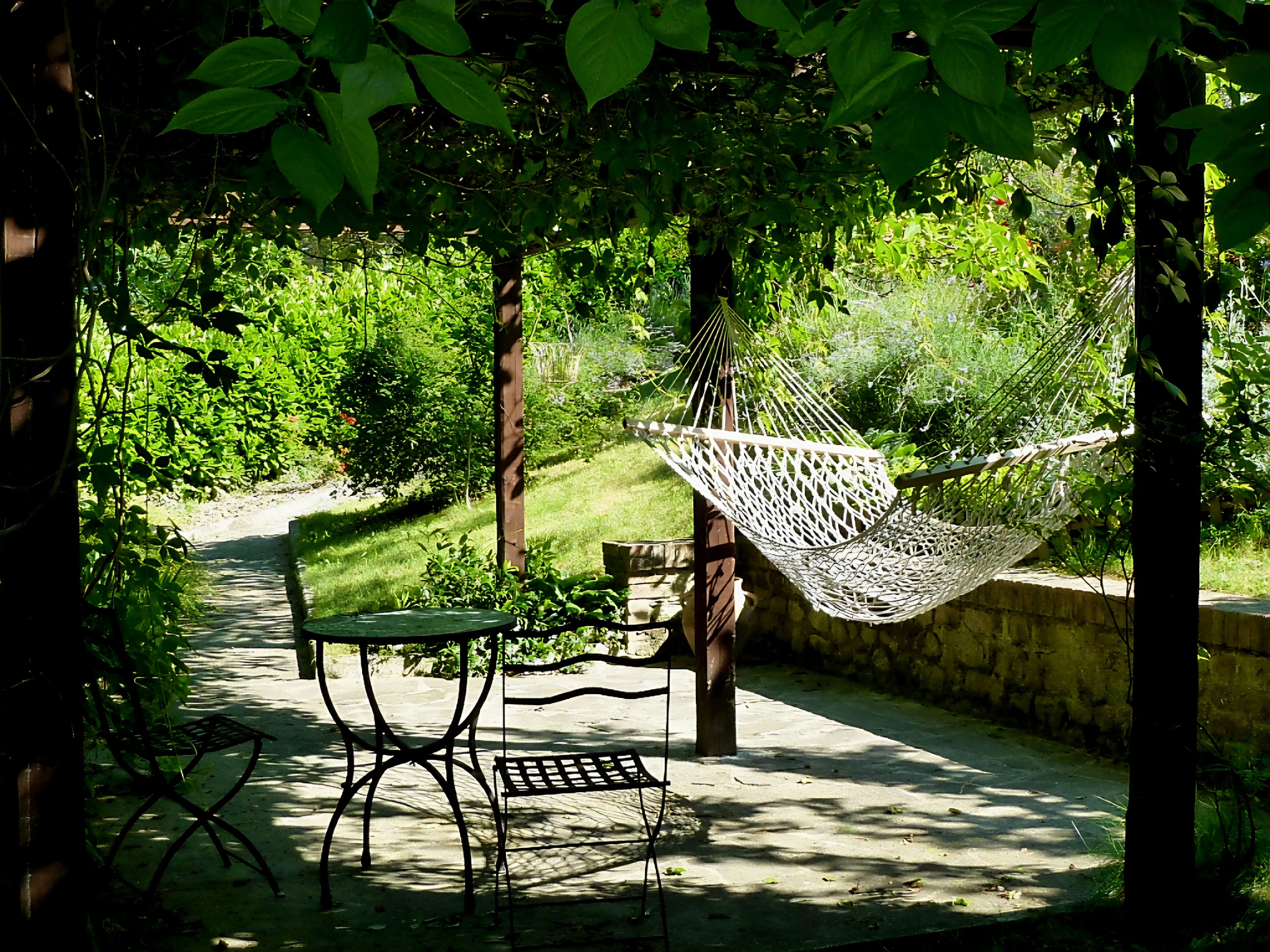 Un hamac, une table en métal et un banc dans un coin de jardin à l’ombre.