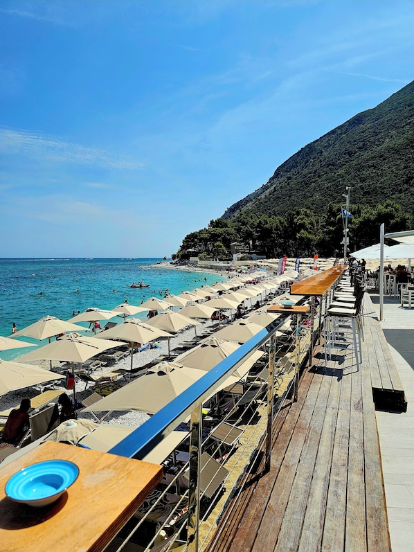 Eine Holzterrasse mit Sitzgelegenheiten und Sonnenschirmen mit Blick auf einen Strand und blaues Wasser sowie eine gebirgige Kulisse.