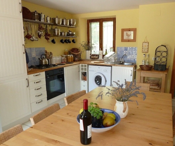 Une cuisine avec des meubles blancs, une table en bois, une bouteille de vin et un saladier rempli de fruits.