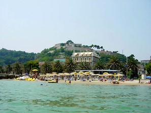 Ein Strand mit Sonnenschirmen, Booten und einer Hügelkette, deren Hintergrund von Gebäuden geprägt ist.