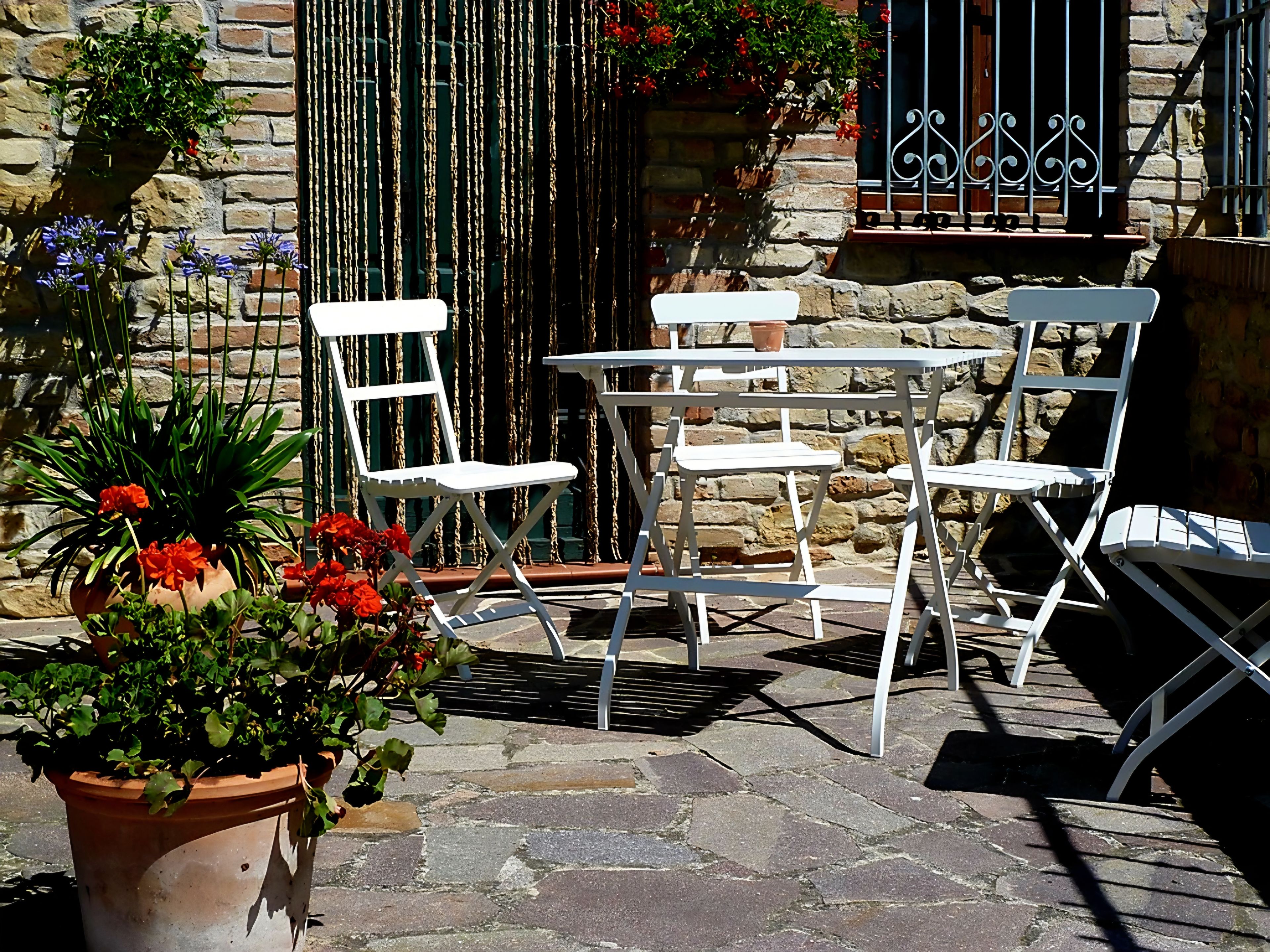 Un área de estar al aire libre con sillas blancas y una mesa, rodeada de plantas en macetas y un muro de piedra.