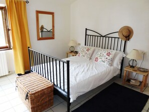 Un dormitorio con una cama, dos mesitas de noche, un tocador, un cofre de mimbre, una ventana con cortinas, un espejo y un sombrero colgado en la pared.