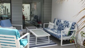 Un espacio exterior moderno con mobiliario blanco, un sofá estampado y un ventanal que da a un ambiente interior.