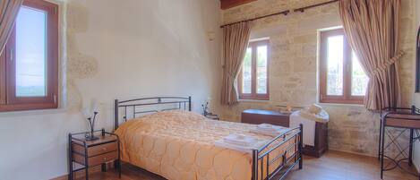 Un dormitorio con techo de madera, una cama con colcha color crema, una mesita de noche con lámpara y una ventana con cortinas.