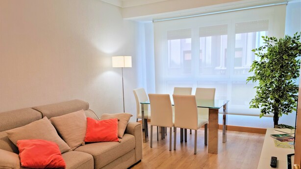 A modern living room with a beige sofa, red cushions, a glass dining table with chairs, a floor lamp, and a potted plant. - EXCLUSIVE APARTMENT SEASIDE BEACH ZURRIOLA (Donostia)