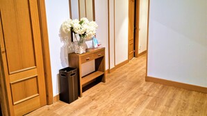 A hallway with wooden flooring, a wooden door, a wooden console table with a mirror, a vase of white flowers, and a trash bin. - EXCLUSIVE APARTMENT SEASIDE BEACH ZURRIOLA (Donostia)