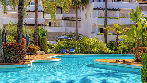 A swimming pool with palm trees, lounge chairs, and a building with balconies in the background. - Puente Romano Phase 2, Golden Mile, Marbella (Marbella)