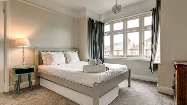 A bedroom with a bed, a bedside lamp, a window with curtains, and a view of buildings outside. - Lavender House | Oxford Holiday Let | 2 Bed & Sleeps 4 (Oxford)