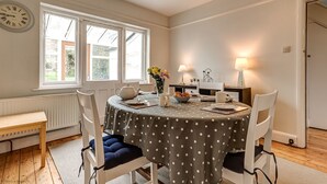 A dining area with a round table covered in a polka dot tablecloth, surrounded by white chairs with blue cushions. A clock is visible on the wall, and a window provides natural light. - Lavender House | Oxford Holiday Let | 2 Bed & Sleeps 4 (Oxford)