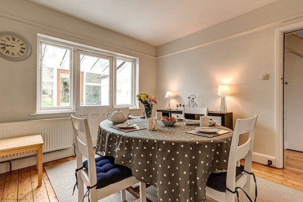 Un coin repas avec une table ronde recouverte d’une nappe à pois, entourée de chaises blanches avec des coussins bleus. Une horloge est visible au mur, et une fenêtre laisse entrer la lumière naturelle.