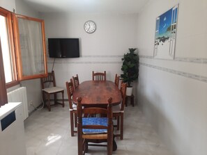 Une salle à manger avec une table en bois, des chaises, une horloge, un téléviseur et une plante en pot.