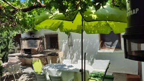 Een terras met een tafel en stoelen onder een groene parasol, een barbecue van baksteen en een stenen muur.