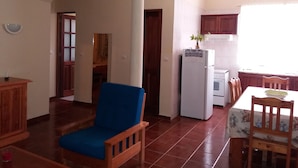 A living room with a blue chair, a dining table with chairs, a kitchen area with a refrigerator, and a window with curtains. - Chã de Arroz Country House (Ribeira Grande)