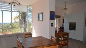 A dining area with a wooden table and chairs, a kitchen with white cabinets and appliances, and a large window with a view of greenery.