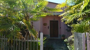 A pink building with a wooden door, surrounded by lush greenery and a white fence.