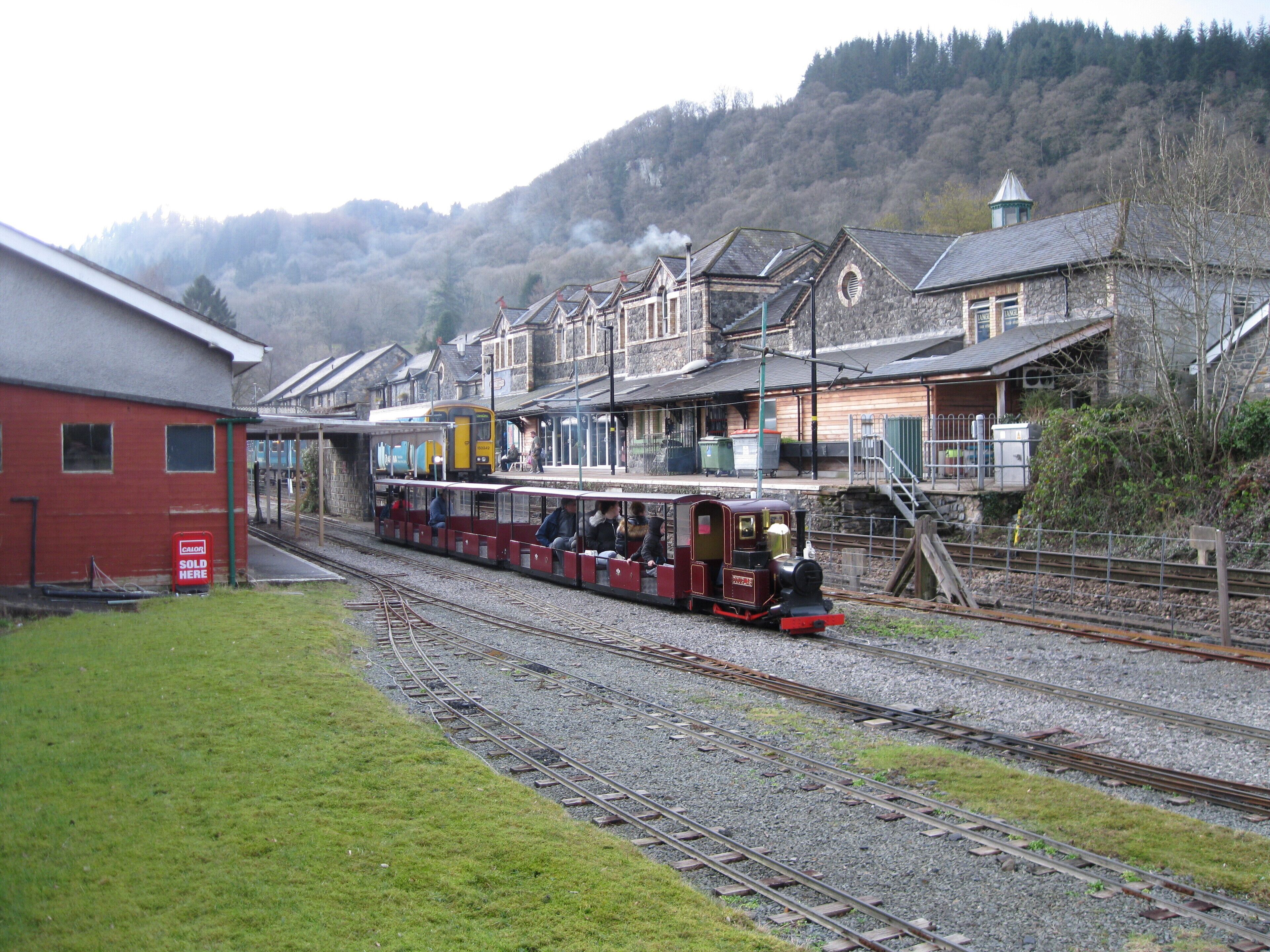 A small train station with a single train on the tracks, surrounded by buildings and a hilly landscape.