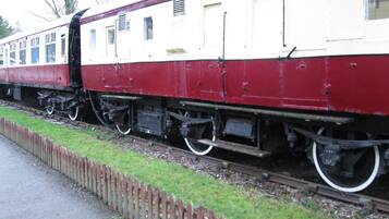 Um trem parado com exterior vermelho e branco, posicionado sobre trilhos ao lado de uma área gramada com cerca de madeira.