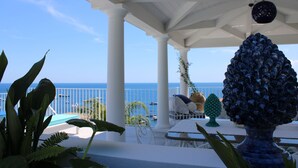 A covered outdoor area with white columns, a table, chairs, and a view of the sea.