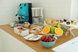 Un comptoir de cuisine avec une machine à café, une cafetière, un bol de fraises, un bol d’oranges et un plateau de viennoiseries.