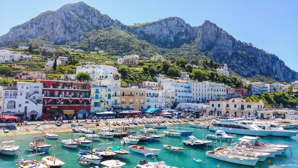 A coastal town with colorful buildings, a marina filled with boats, and a mountainous backdrop.