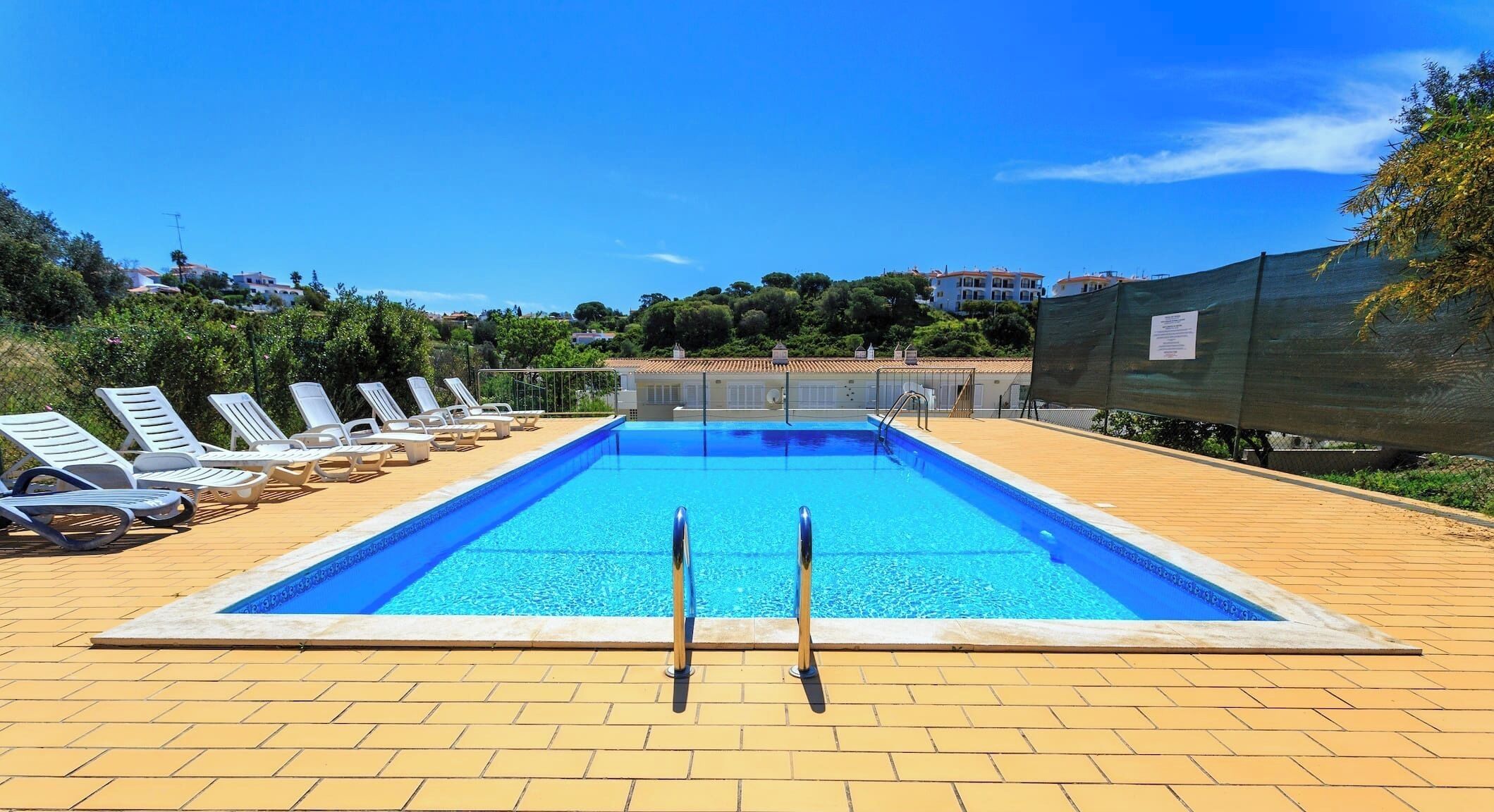 A rectangular swimming pool with two ladders, surrounded by sun loungers and a tiled deck.