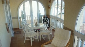 A sunlit patio with a round table and white chairs, a cushioned chair, and a view through a glass door. - Villa with Private Pool and Large Underbuild (Gran Alacant)