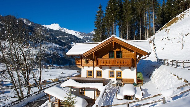 Una cabaña de madera con balcón, rodeada de montañas y árboles cubiertos de nieve.