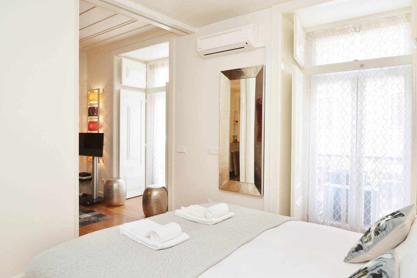 A hotel room with a bed, a flat-screen TV, a mirror, and a window with sheer curtains.