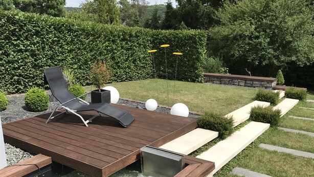 Une terrasse en bois avec un jacuzzi, entourée d’un jardin agrémenté de haies et d’un mur en pierre.