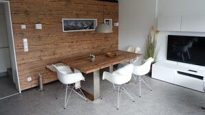 Un espace repas moderne avec un mur en bois, des chaises blanches, une table en bois et un téléviseur à écran plat fixé au mur.