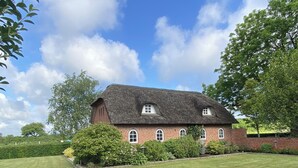 Een bakstenen huis met rieten dak, omgeven door een verzorgd gazon en volwassen bomen.