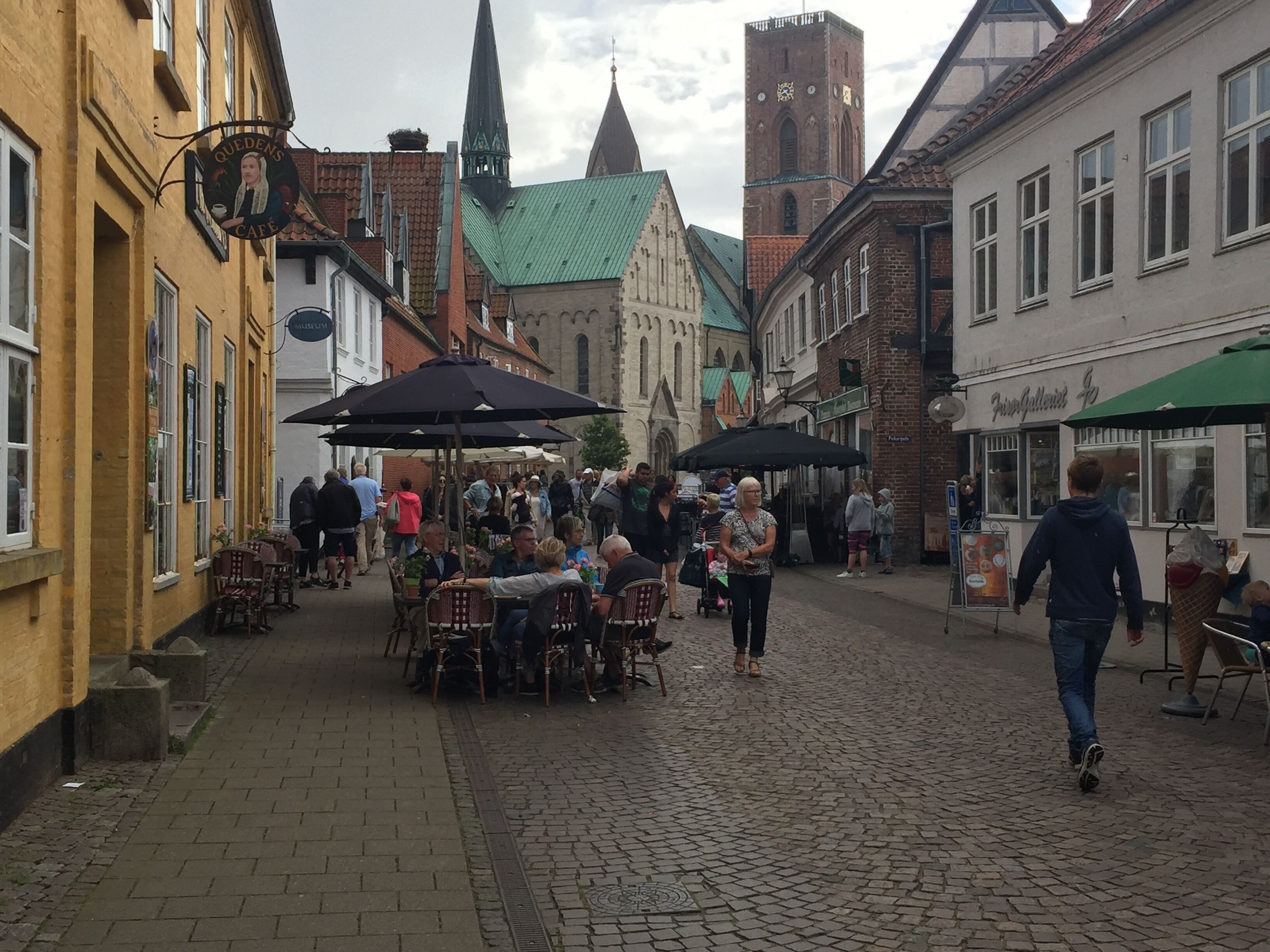 Une rue pavée, avec des tables et des chaises installées à l’extérieur et des parasols, bordée de bâtiments historiques et dominée par le clocher d’une église.