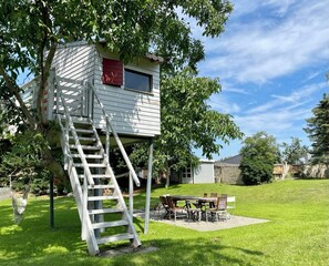 Una casa en un árbol con una escalera de madera, una mesa de picnic y sillas en un patio trasero.