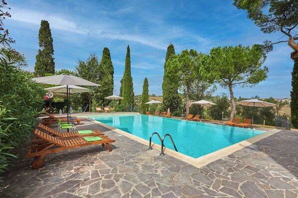 A swimming pool with lounge chairs and umbrellas, surrounded by trees and stone flooring.