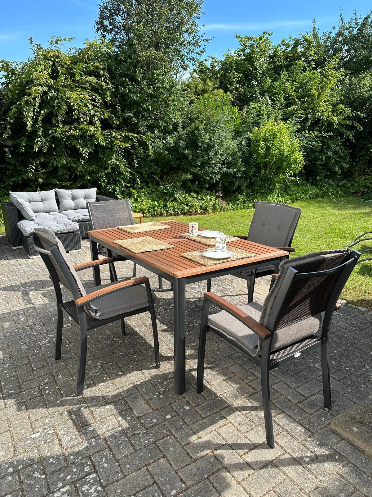 Een terras buiten met een houten tafel en stoelen, omringd door weelderige begroeiing.
