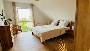 Une chambre avec un plancher en bois, un lit recouvert de draps blancs, une table de chevet, un luminaire suspendu, une fenêtre avec des rideaux et une vue sur la verdure à l’extérieur.