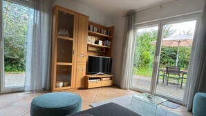 Un salon moderne avec un canapé, un téléviseur et une bibliothèque. Une porte-fenêtre coulissante donne accès à un espace extérieur aménagé avec une table et des chaises.