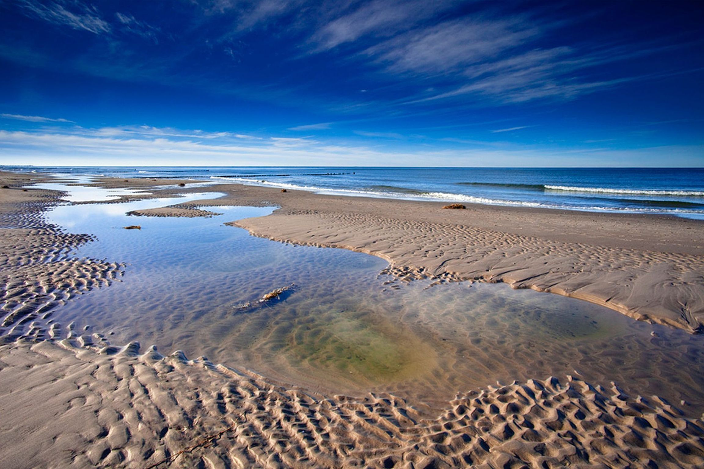 Ein Strand mit klarem Wasser, sichtbaren Fußspuren und einer fernen Küstenlinie.