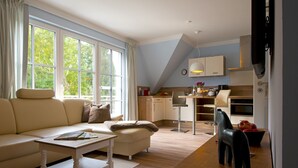 Ein modernes Wohnzimmer mit Sofa, Couchtisch und Essbereich mit Stuhl und Tisch. Im Hintergrund ist eine Küche mit Schränken und Geräten zu sehen.