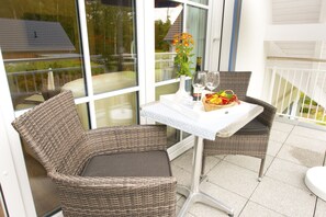 Un balcon avec des fauteuils en osier, une table recouverte d’une nappe blanche, un vase rempli de fleurs et un panier de fruits.