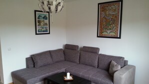 Ein Wohnzimmer mit einem grauen Ecksofa, einem schwarzen Couchtisch mit einer Kerze, einem gemusterten Teppich und zwei gerahmten Kunstwerken an den Wänden.