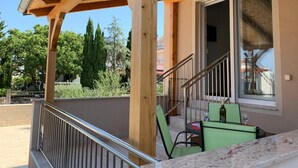 Un balcon en bois avec une rambarde en métal, un escalier menant à l’étage supérieur et, au loin, une vue sur les arbres et les bâtiments.