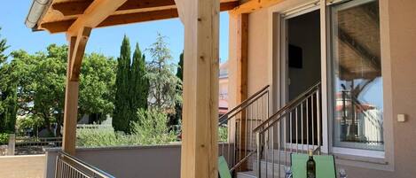 A wooden balcony with a metal railing, a staircase leading to an upper floor, and a view of trees and buildings in the background.