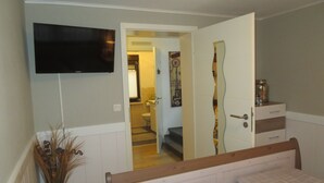 A bedroom with a bed, a TV mounted on the wall, a door leading to a bathroom, and a small cabinet with a jar. - Vacation apartment Marie in Bergen auf Rügen, quiet location (Bergen auf Rügen)