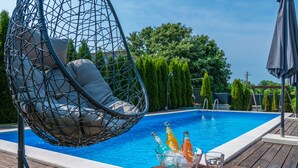 A poolside area with a hanging chair, a wooden table with bottles and glasses, and a clear blue pool surrounded by greenery.